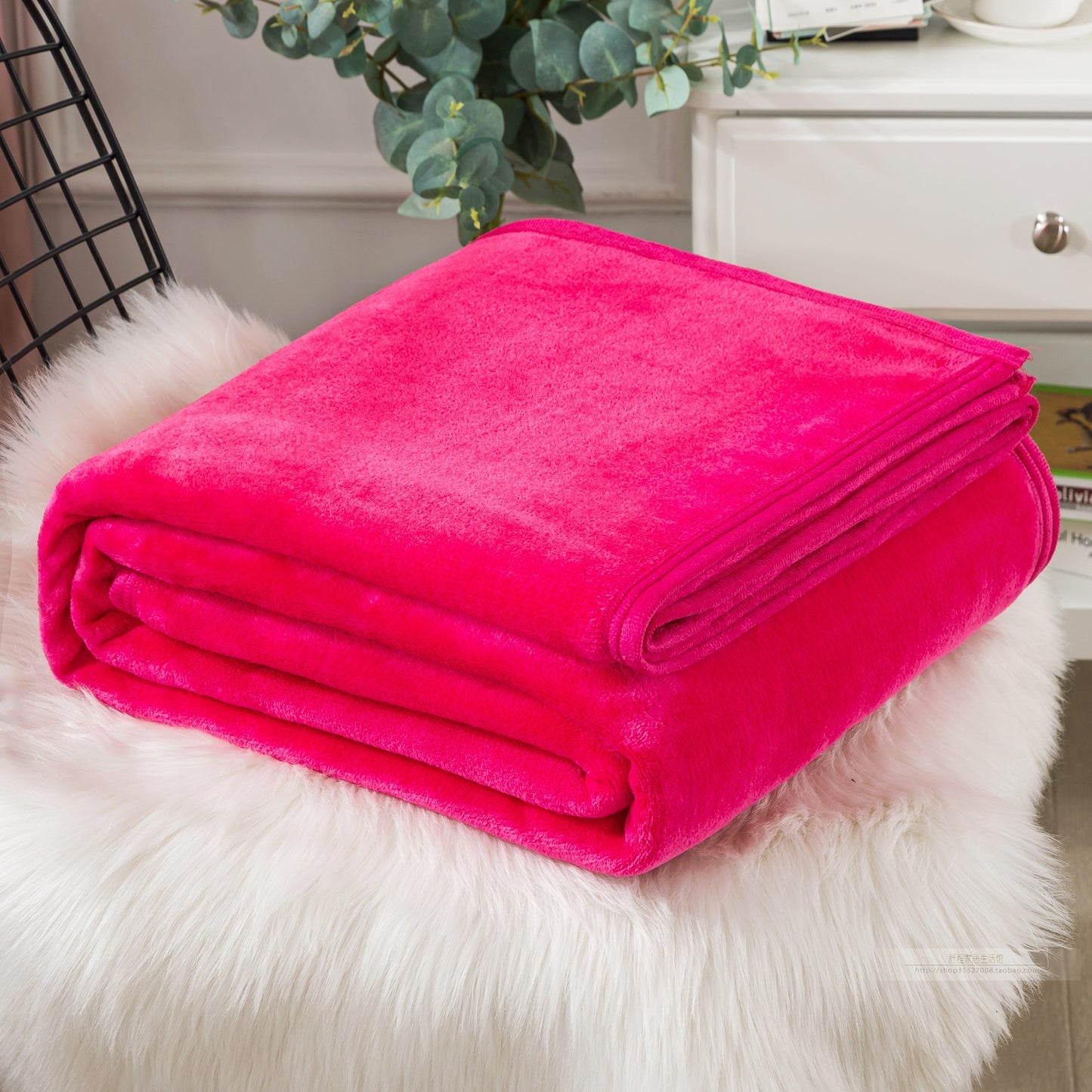 Folded pink blanket on a white fur surface with a decorative plant and cabinet in the background.