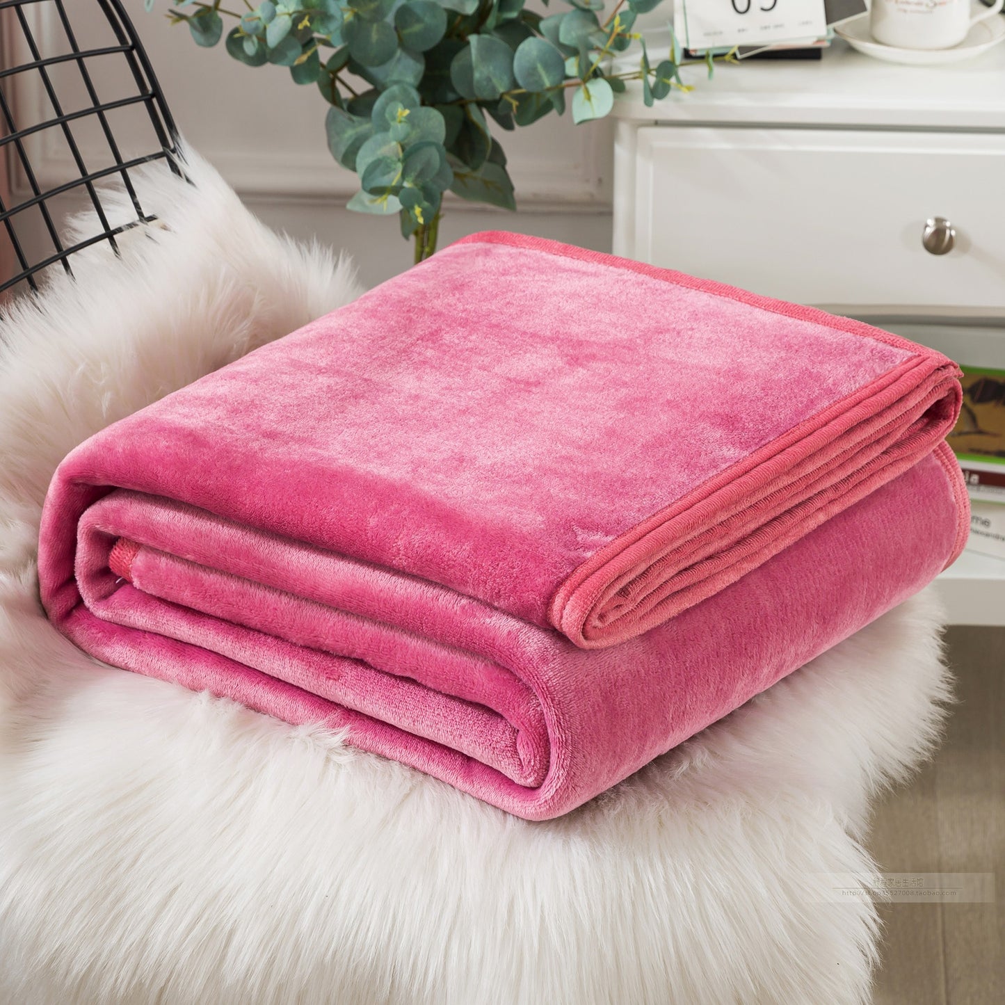 A blue coral fleece blanket placed on a white furry surface, with a part of a bed and a plant in the background.
