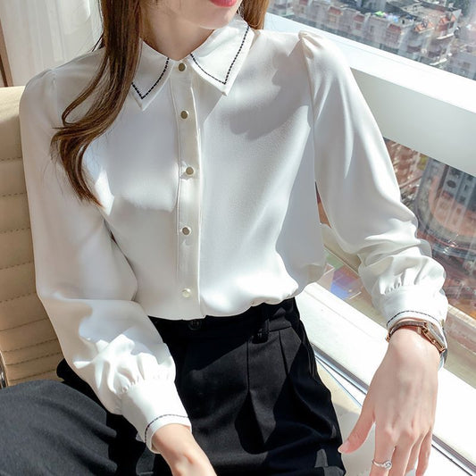 Woman wearing a white blouse with black trim sitting by a window with a cityscape view.