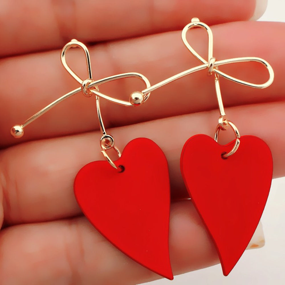 Red heart-shaped earrings with gold bows held between fingers