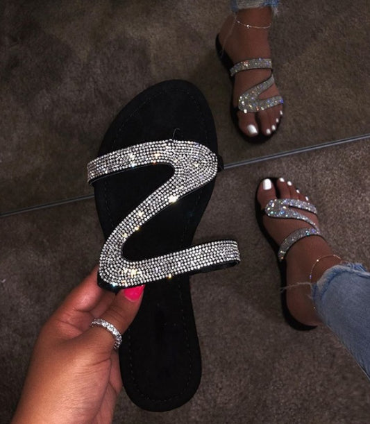 Black sandal with rhinestone strap held by a hand on a tiled floor.