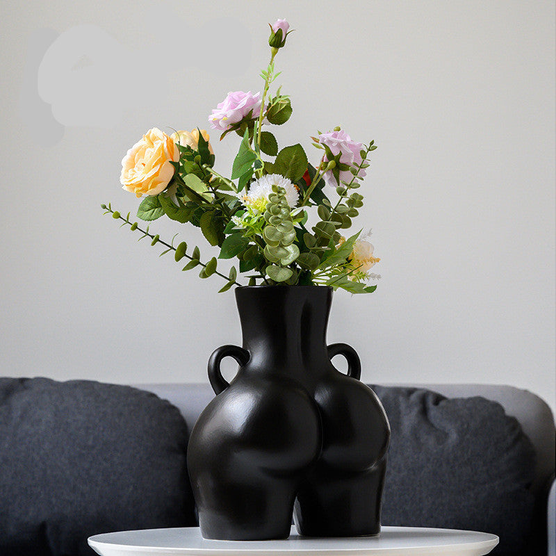 A black ceramic gourd-shaped vase on a white surface, with a bouquet of multicolored flowers arranged in it, placed against a neutral background with a grey sofa.