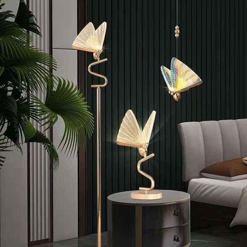 A modern floor lamp with a butterfly design, featuring gold-colored metal bodies and multicolored acrylic shades, standing in a room with a dark wall and a plant.