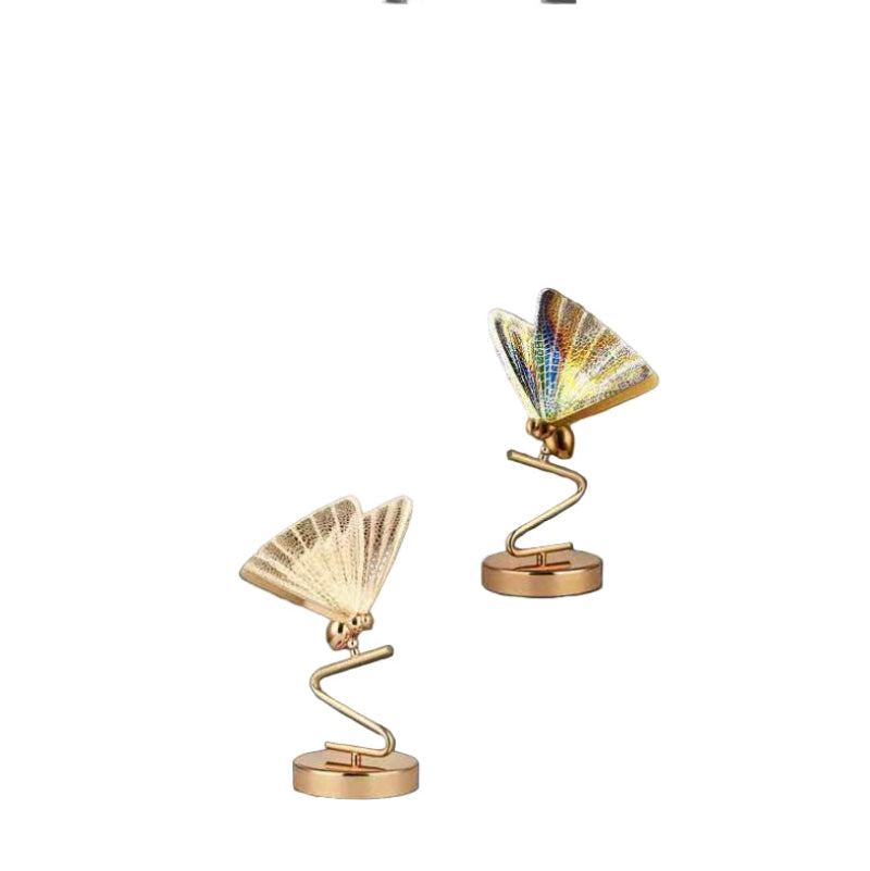 A modern floor lamp with a butterfly design, featuring gold-colored metal bodies and multicolored acrylic shades, standing in a room with a dark wall and a plant.