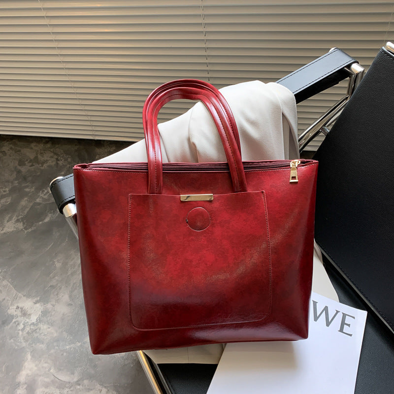 A beige tote bag with a zipper and soft handles placed on a chair, with a white shirt and a black bag in the background.