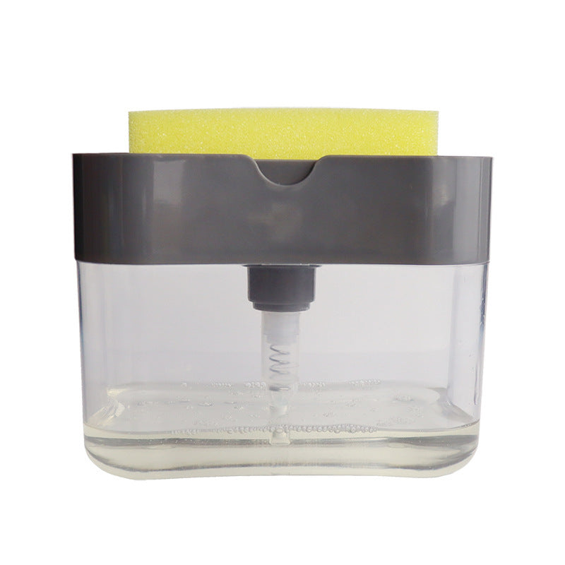 A person's hand pressing down on a sponge attached to a clear soap dispenser caddy, which is mounted on a wall beside a sink.