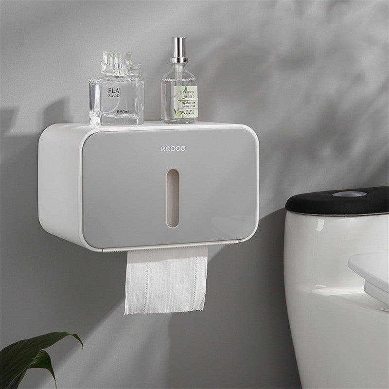 White paper towel holder mounted on a wall with two bottles and a plant in the background.