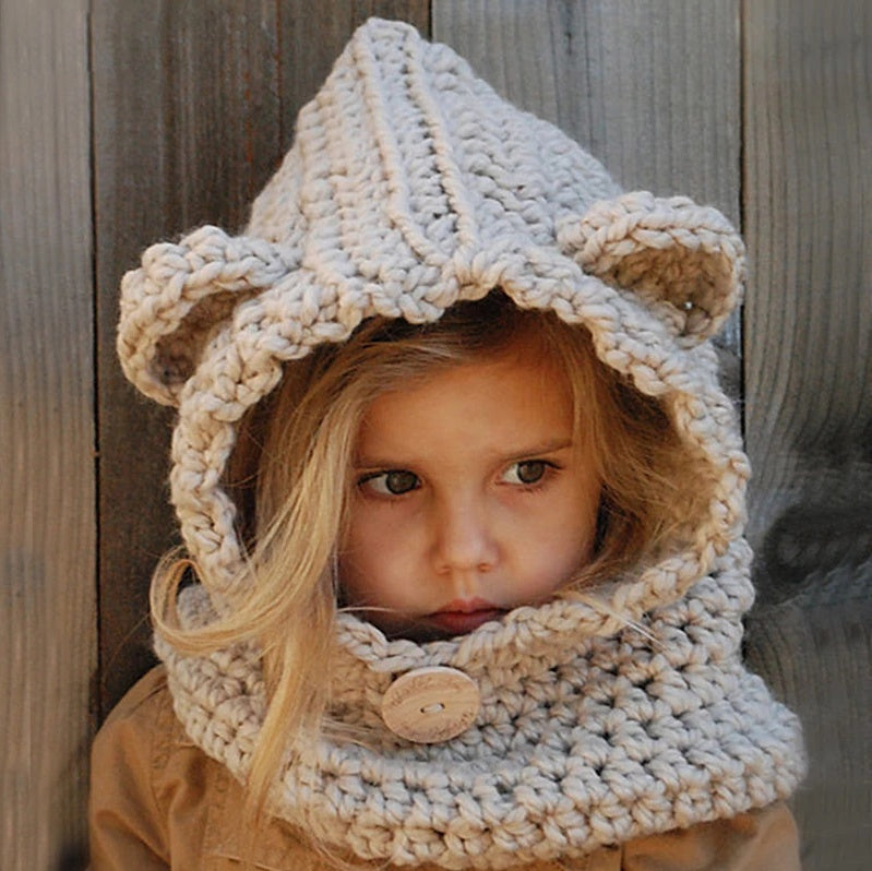 A baby wearing a beige knitted hat with a hood that has a bear face design and wooden button details.