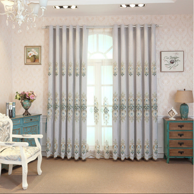 A room with light blue embroidered curtains hanging on a window, with a white chair and a lamp in the foreground.