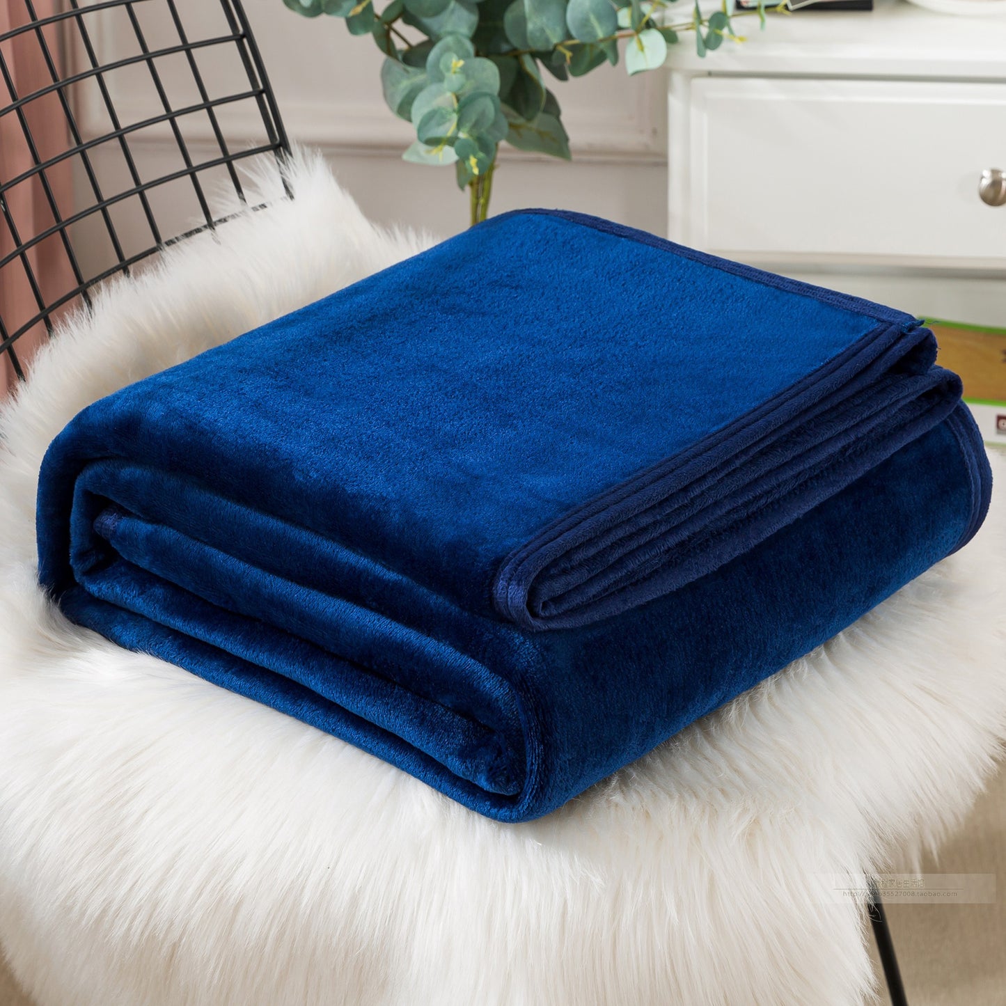A blue coral fleece blanket placed on a white furry surface, with a part of a bed and a plant in the background.