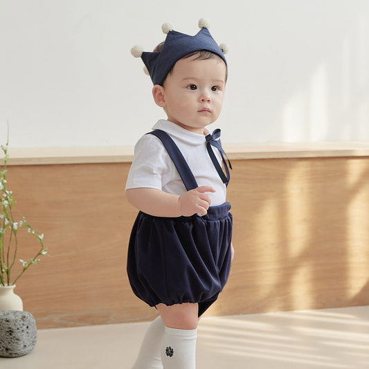 Baby wearing a navy outfit with suspenders and a crown, standing indoors.