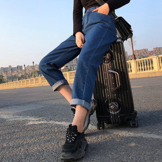 Person wearing blue jeans and black shoes standing next to a black suitcase on a street.