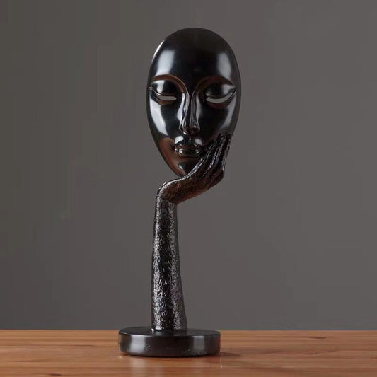A black sculpture of a stylized face with a finger to the lips, placed on a wooden surface against a neutral background.