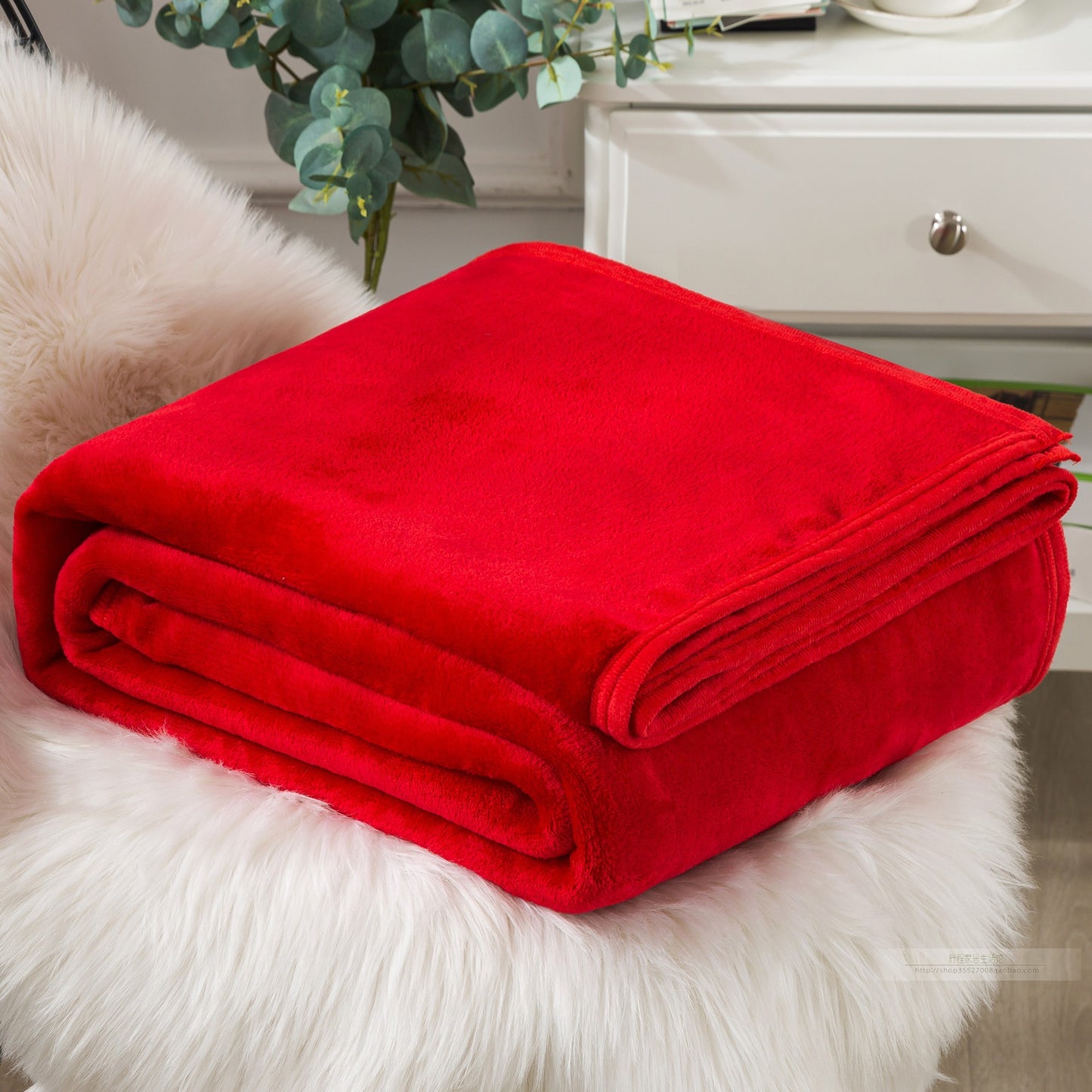 A blue coral fleece blanket placed on a white furry surface, with a part of a bed and a plant in the background.