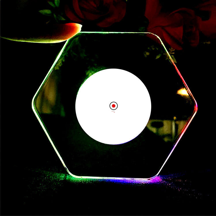 An LED glow coaster with multicolored lights around its edge, placed on a glass with ice cubes and a green garnish on top, on a dark background.
