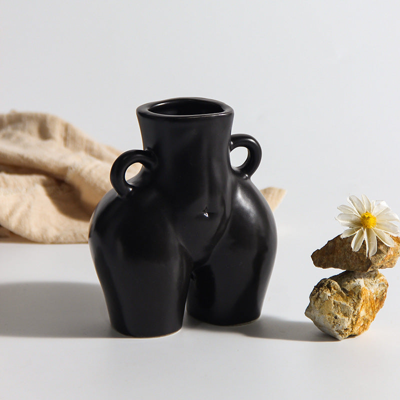 A black ceramic gourd-shaped vase on a white surface, with a bouquet of multicolored flowers arranged in it, placed against a neutral background with a grey sofa.