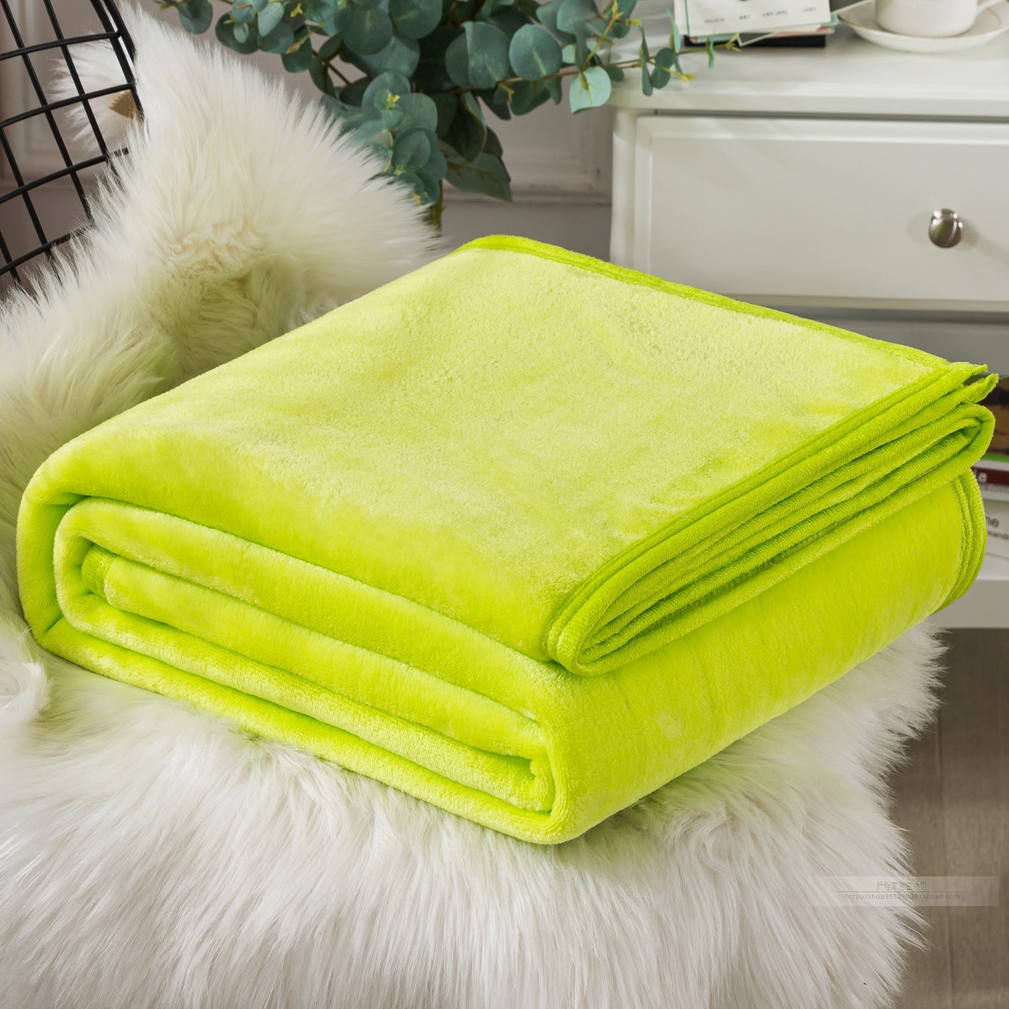 A blue coral fleece blanket placed on a white furry surface, with a part of a bed and a plant in the background.