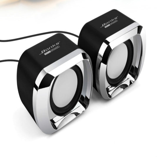 Two portable USB mini speakers on a wooden desktop surface, one black and one white, with a potted plant and a laptop in the background.