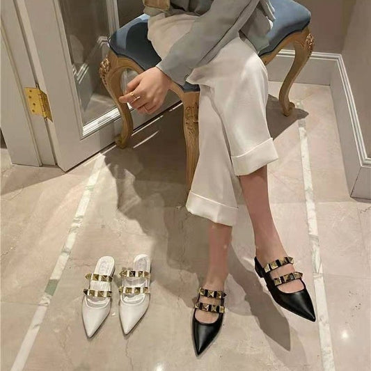 Person sitting on a chair wearing black and white pointed-toe shoes with gold buckles, with shoes beside them on the floor.