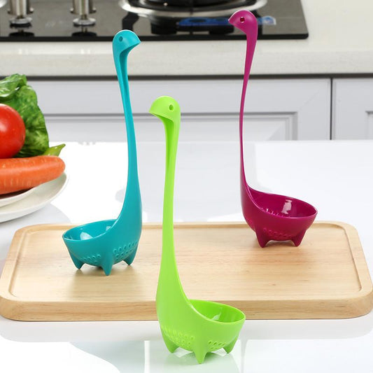 Three colorful soup spoons designed to resemble dinosaurs, in green, blue, and rose colors, standing upright on a wooden board in a kitchen setting.