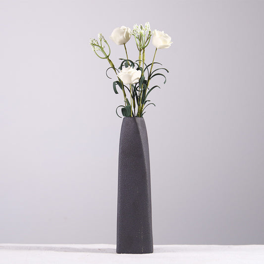 A black, ceramic, cylindrical vase with a narrow top, standing on a flat surface, with a single white flower inside it.