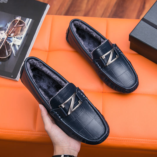 Black loafers with a brand logo held by a hand on an orange surface.