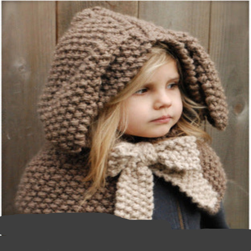 A baby wearing a beige knitted hat with a hood that has a bear face design and wooden button details.