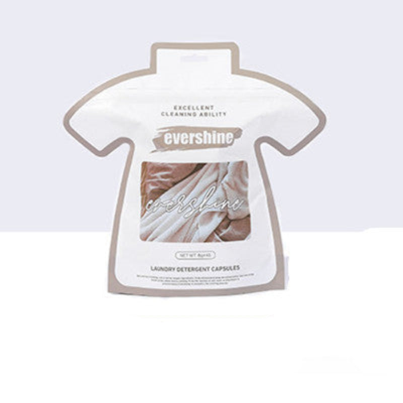 Packaging of laundry gel beads in the shape of a shirt placed on a washing machine, with plants and laundry in the background.