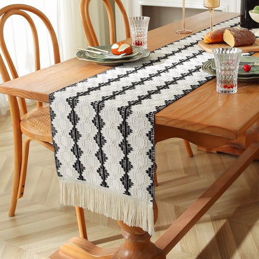 A rectangular tablecloth with a geometric pattern, primarily in black and white, laid out on a wooden dining table. The tablecloth features a tassel detail at the end.