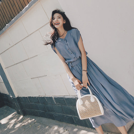 Woman in a blue dress holding a woven handbag against a white wall.