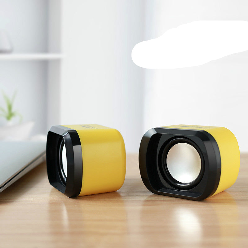 Two portable USB mini speakers on a wooden desktop surface, one black and one white, with a potted plant and a laptop in the background.
