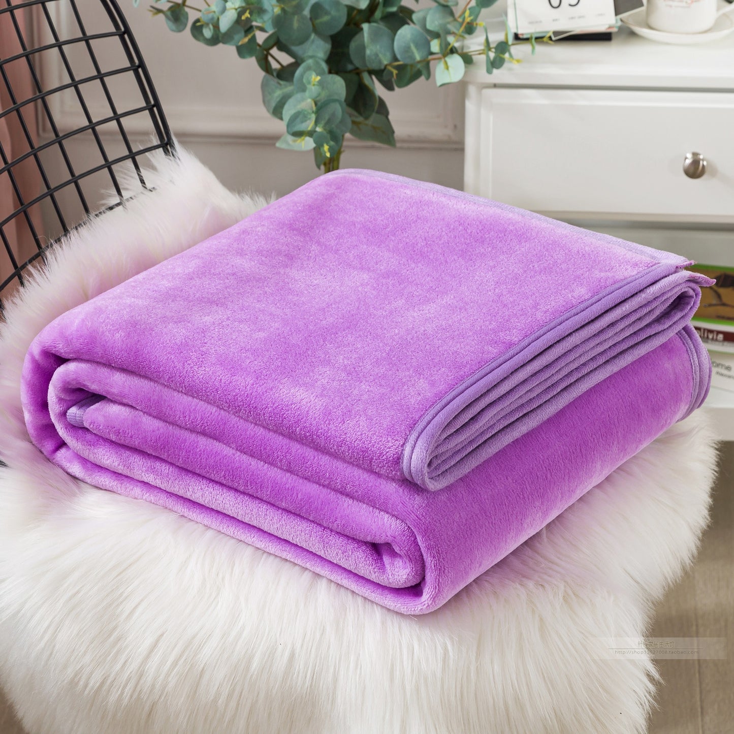 A blue coral fleece blanket placed on a white furry surface, with a part of a bed and a plant in the background.