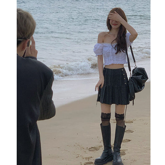 Woman posing on a beach with a person taking a photo, wearing a white top and black pleated skirt.