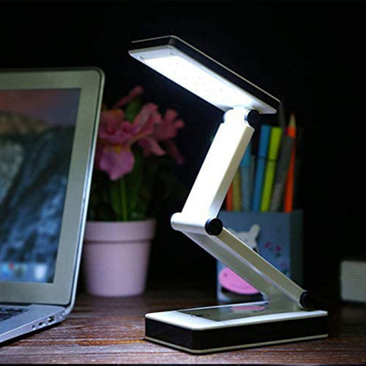 A foldable desk lamp with a white and black color scheme, illuminated and placed on a desk next to a laptop and pens, with a dimmer switch on the base.