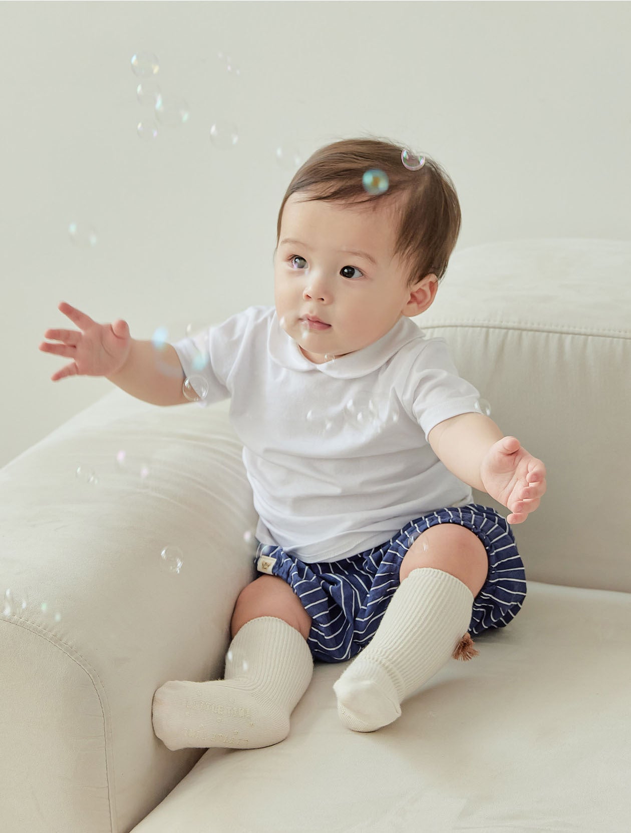 Baby sitting on a white couch wearing a white shirt and blue shorts with white socks.