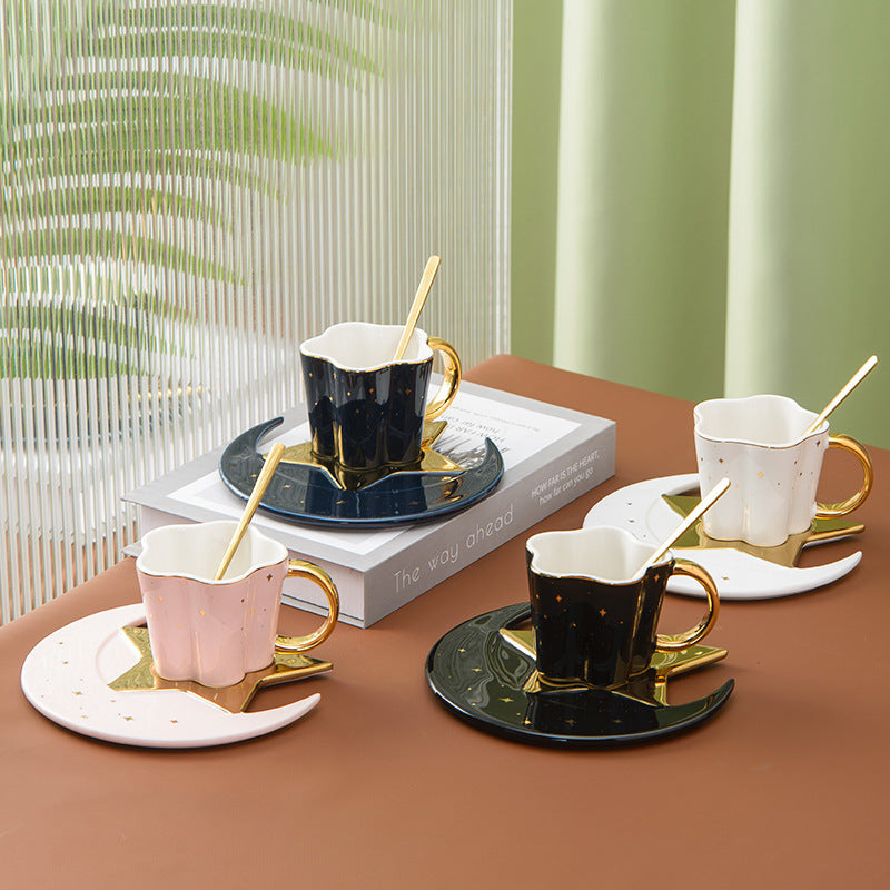 A set of ceramic cups with star and moon saucers in black, white, and gold colors, displayed on a table with books and decorative stars.
