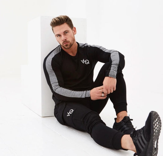 Man wearing a black and gray tracksuit with a logo, sitting on a white surface.