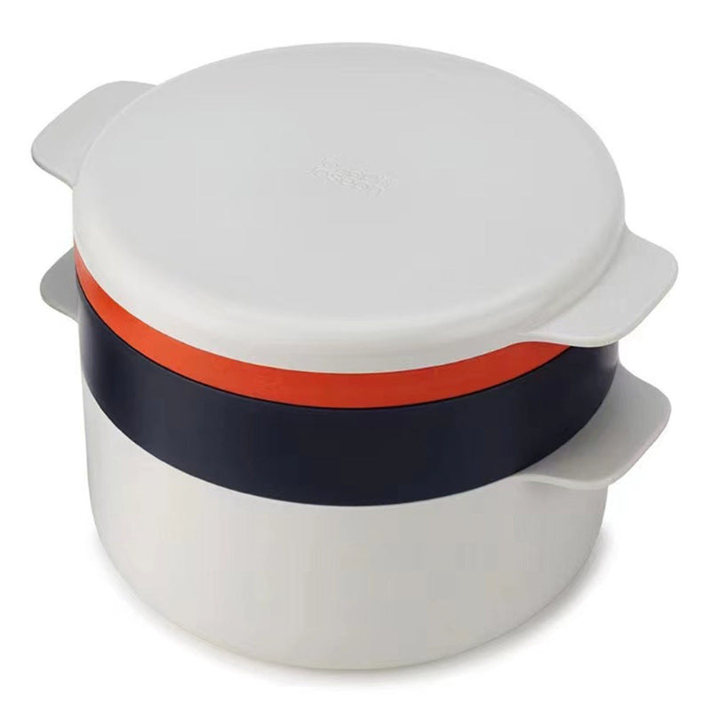 A white, gray, and orange kitchen plastic microwave steaming container with food inside, placed on a table with utensils and plates of food in the background.