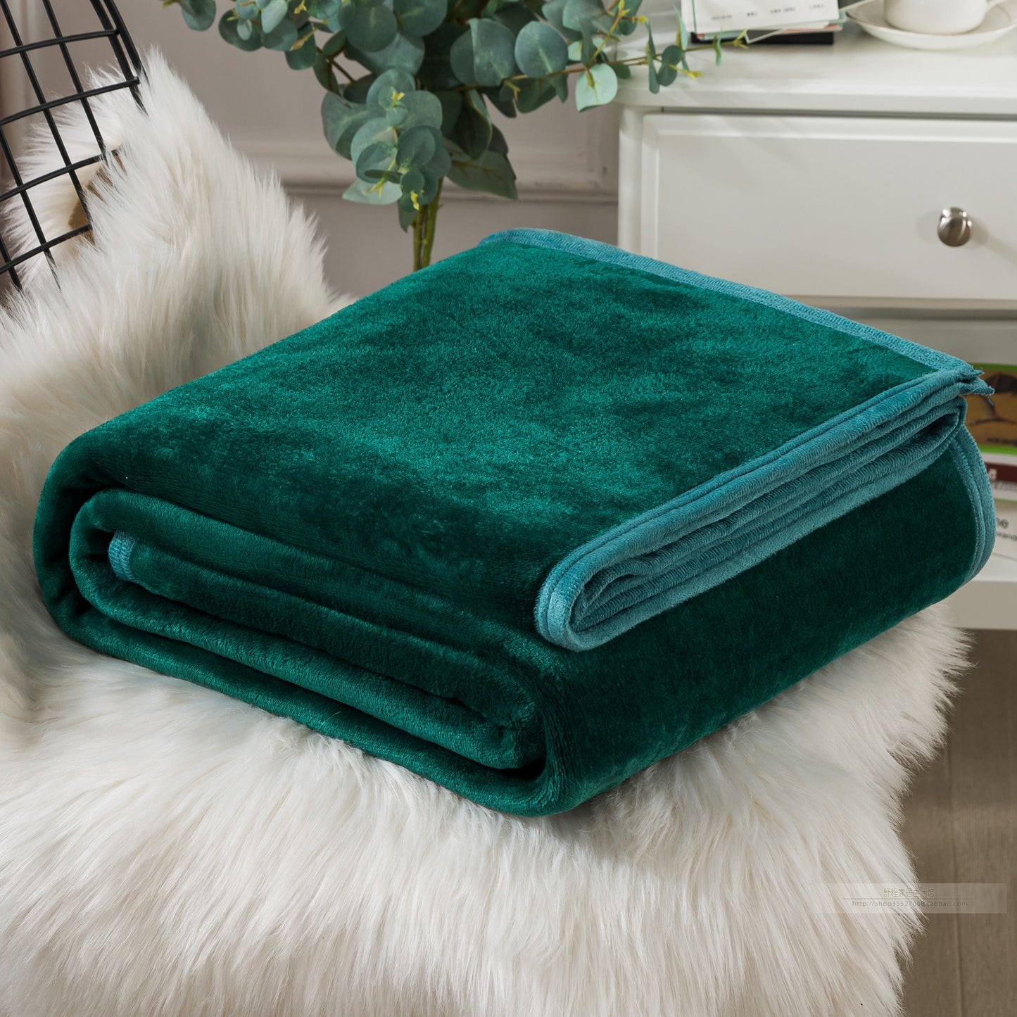 A blue coral fleece blanket placed on a white furry surface, with a part of a bed and a plant in the background.