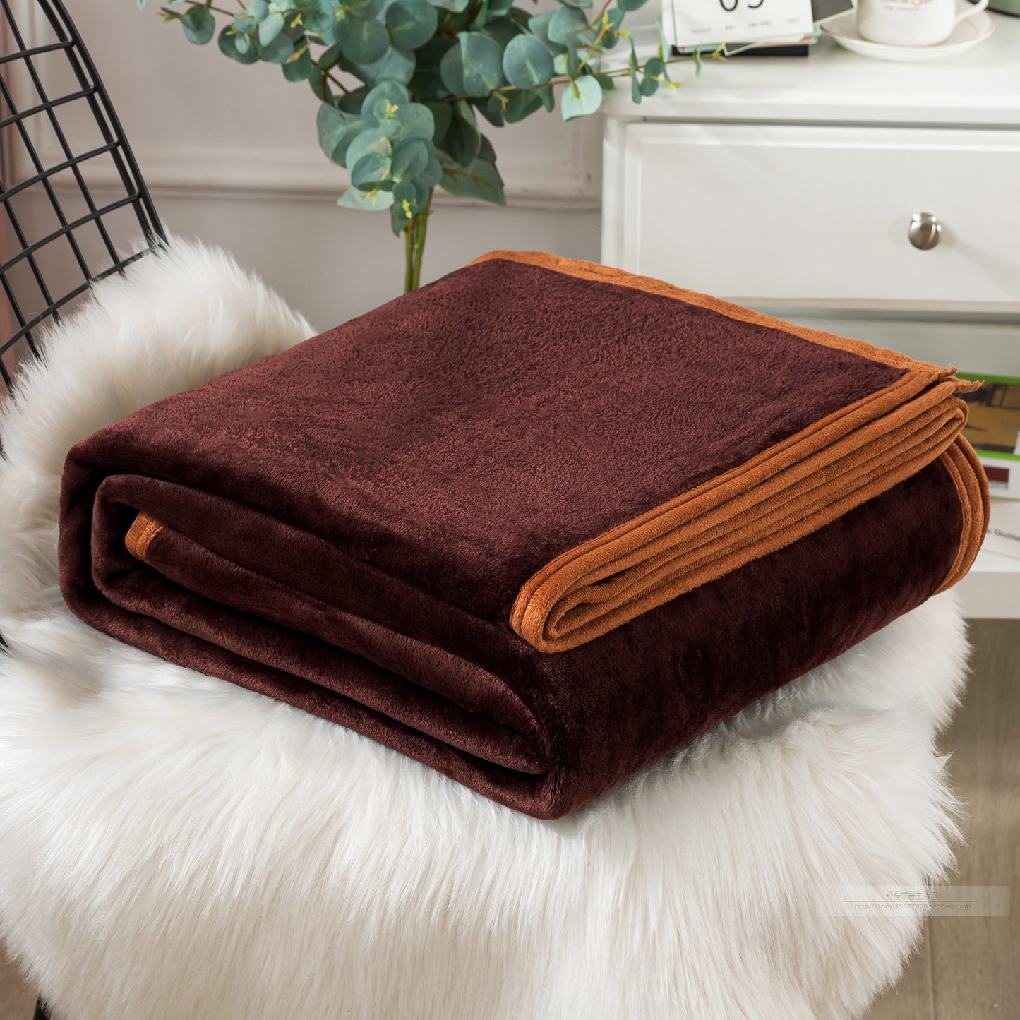 A blue coral fleece blanket placed on a white furry surface, with a part of a bed and a plant in the background.