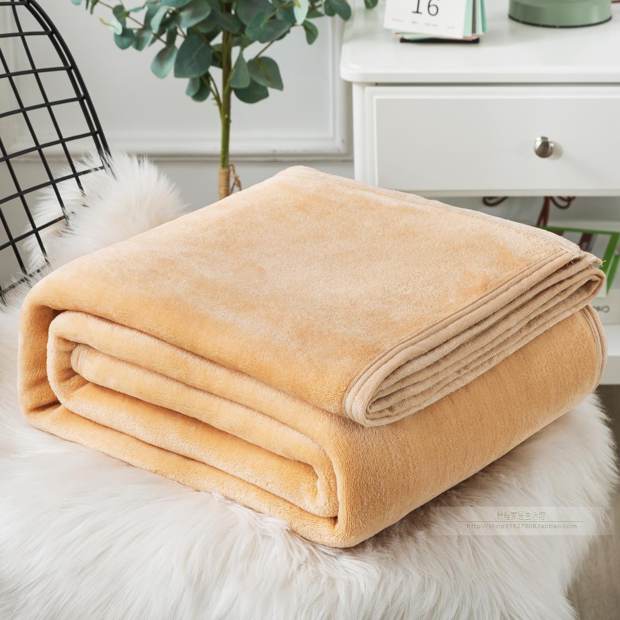 A blue coral fleece blanket placed on a white furry surface, with a part of a bed and a plant in the background.