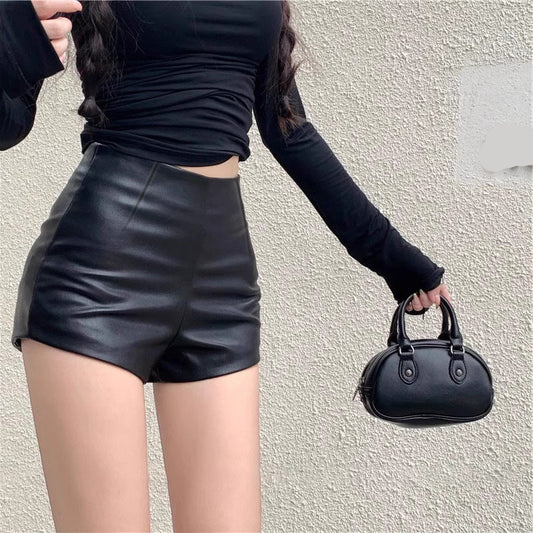 Person wearing black leather shorts and holding a matching black handbag against a light-colored wall.