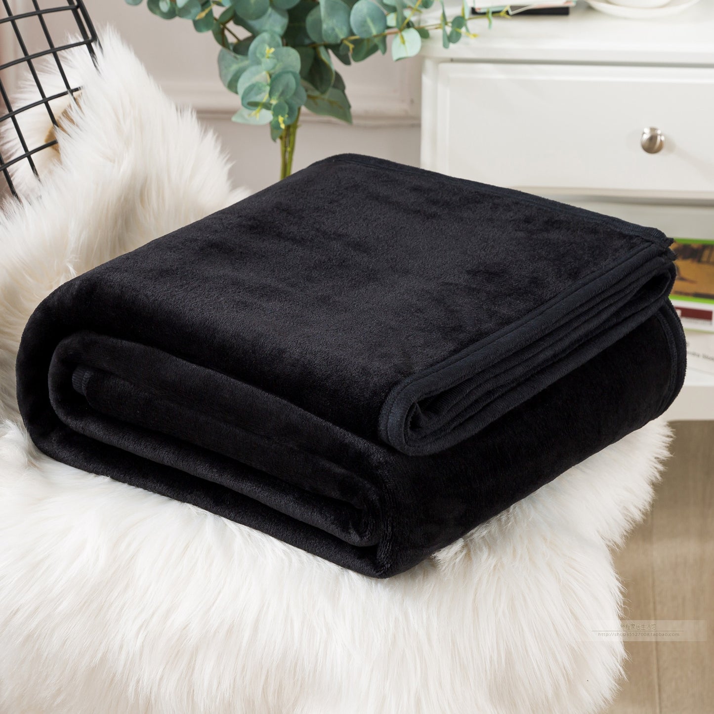 A blue coral fleece blanket placed on a white furry surface, with a part of a bed and a plant in the background.