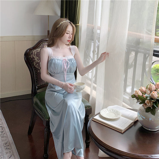 Woman in a light blue dress sitting by a window with a cup, surrounded by books and flowers.