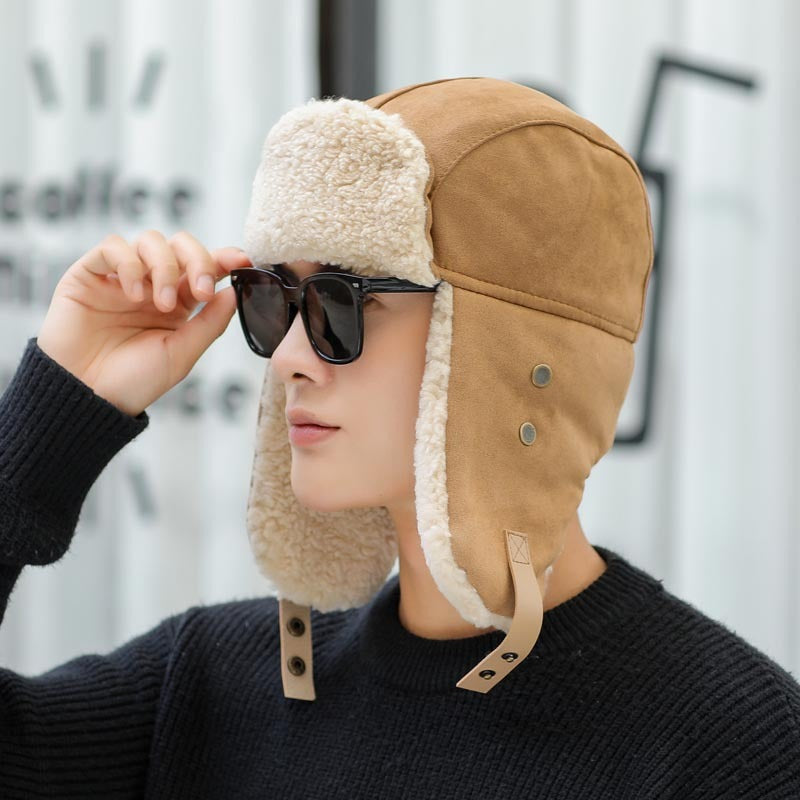 Person wearing a tan aviator hat with ear flaps and holding sunglasses, against a blurred background.