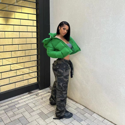 Person wearing a green puffer jacket and camouflage pants standing against a wall.