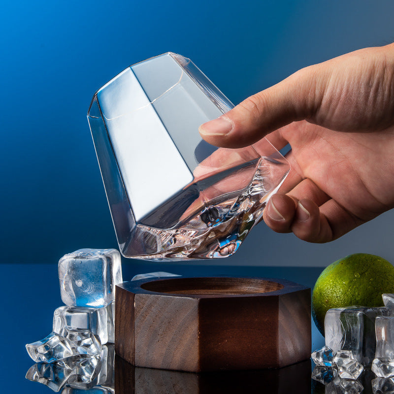 A hand holding a clear crystal wine glass with a wooden round base on a table, with ice cubes and a lime in the background.