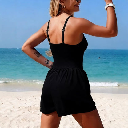 Woman in a black dress standing on a beach with clear blue water and sky.