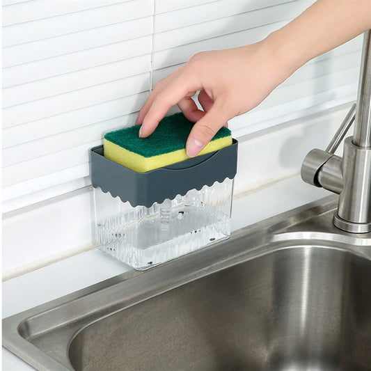 A person's hand pressing down on a sponge attached to a clear soap dispenser caddy, which is mounted on a wall beside a sink.