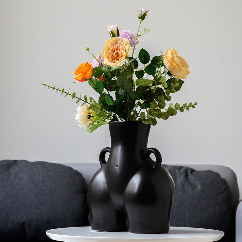 A black ceramic gourd-shaped vase on a white surface, with a bouquet of multicolored flowers arranged in it, placed against a neutral background with a grey sofa.